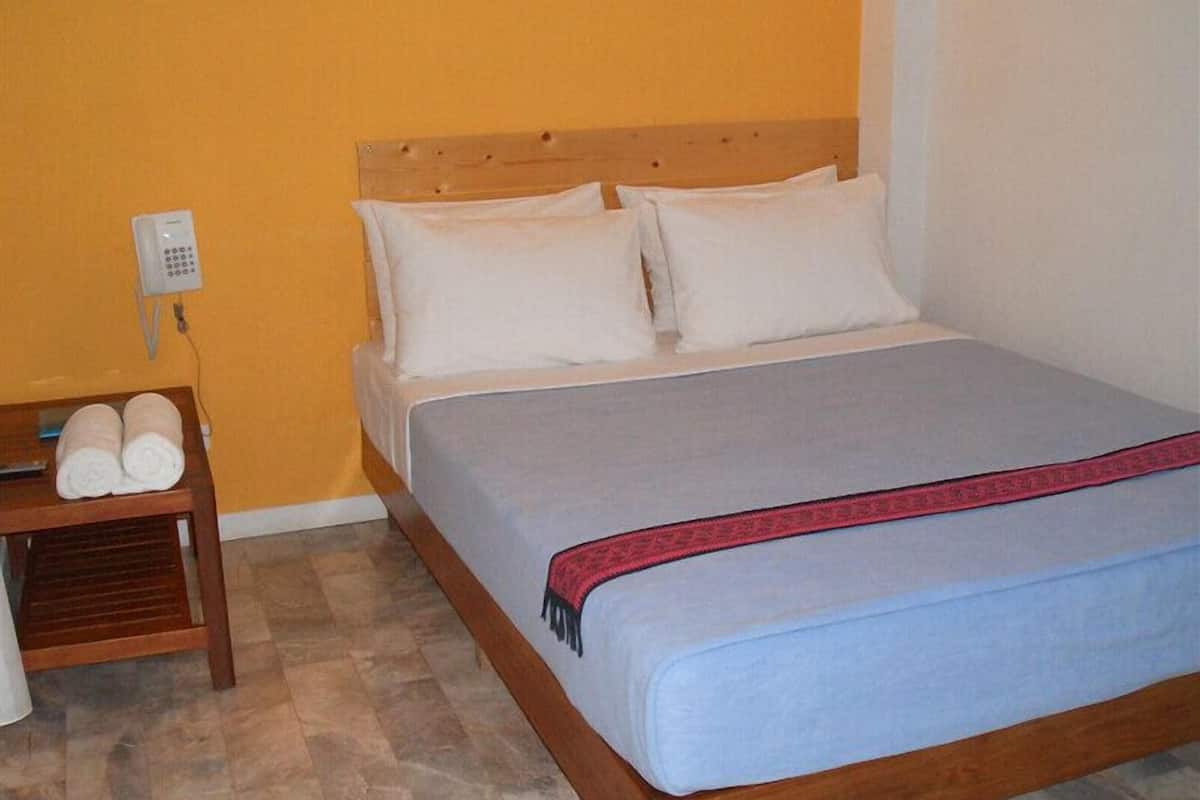 A single bed with a wooden headboard, white bedding, and a red blanket. There is a wooden nightstand with towels and a telephone.