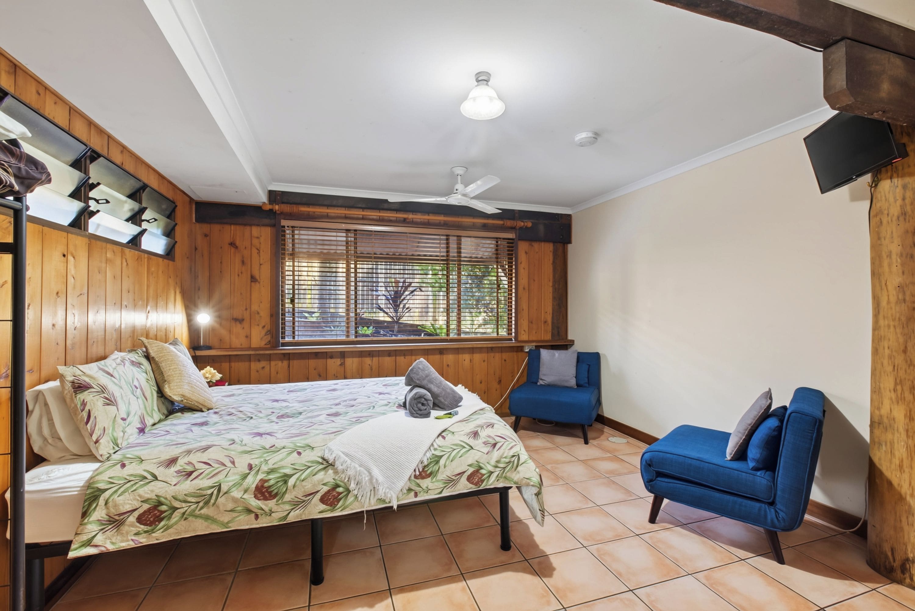 Une chambre à coucher avec un lit, une chaise, une fenêtre avec des stores et un ventilateur de plafond.
