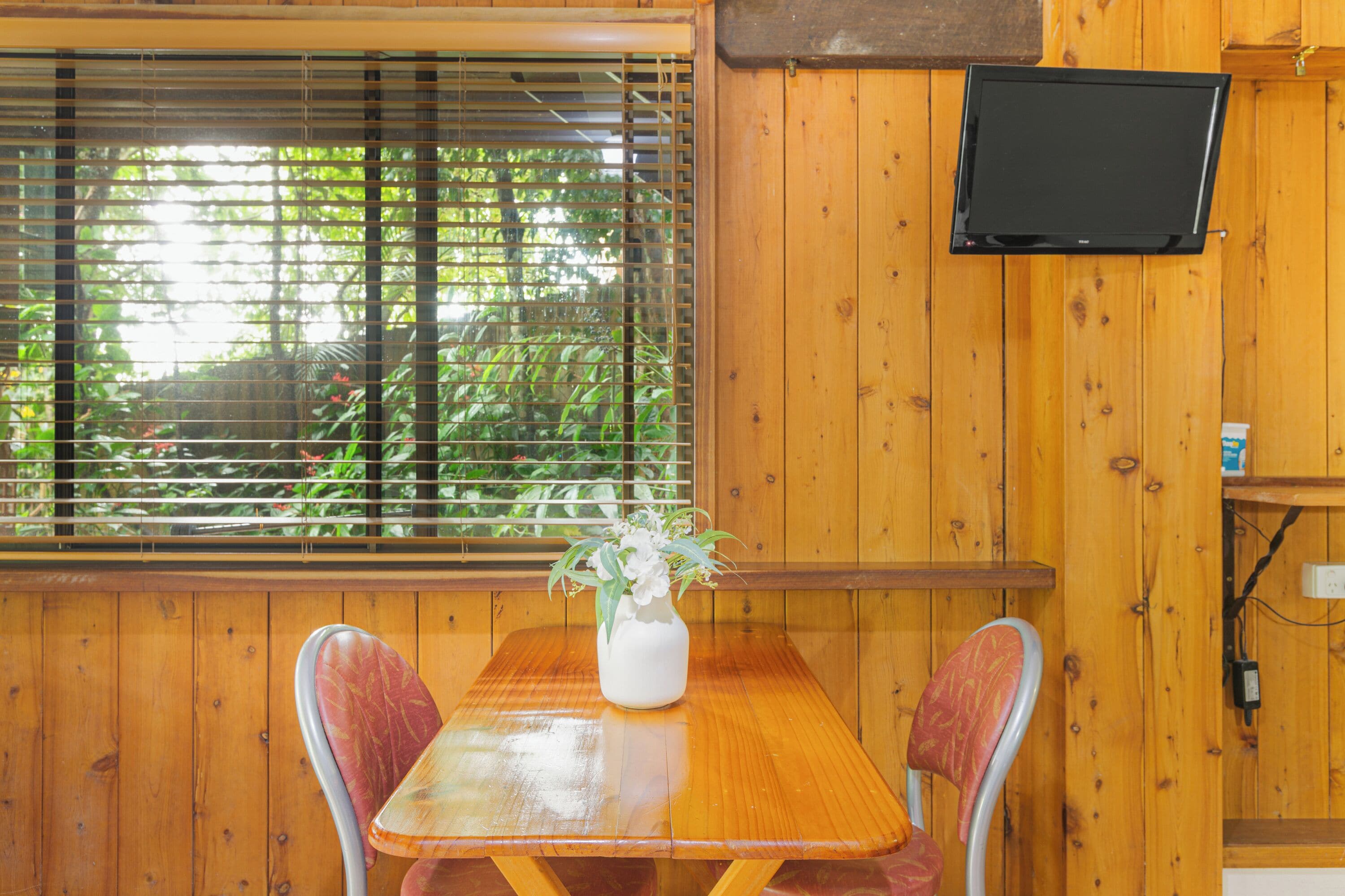 A wooden interior with a table, chairs, a TV mounted on the wall, and a window with blinds.