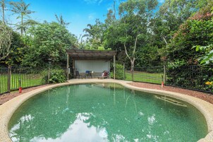 Una piscina cristalina con cubierta de madera y zona de estar, rodeada por una valla y abundante vegetación.