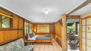 Een houten kamer met een bed, een bank en een tafel. Er is een raam met zonwering en een deur naar een buitengebied.
