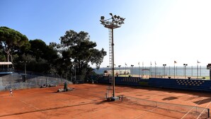 A tennis court with a tall floodlight, a net, and a seating area with chairs. - Hotel Petit Royal (Ospedaletti)
