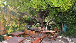 An outdoor seating area with wooden tables and chairs, surrounded by lush greenery and a large tree. - Hotel Petit Royal (Ospedaletti)