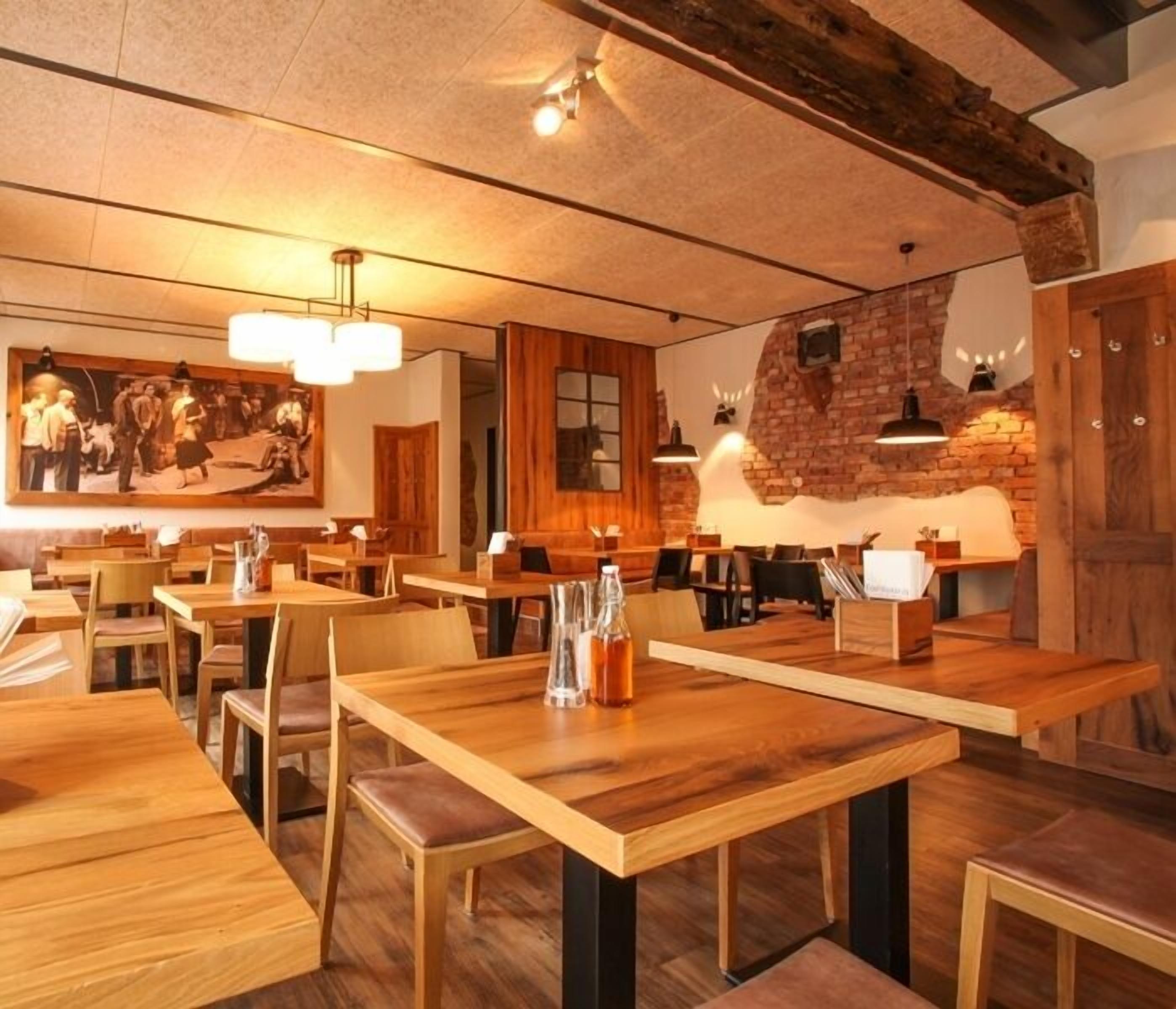 A restaurant interior with wooden tables and chairs, a brick wall, and hanging pendant lights.