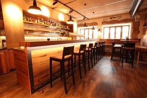 A bar area with wooden bar counter, multiple bar stools, shelves with bottles and glasses, and a brick wall with windows. - Hotel Kunstmühle (Mindelheim)
