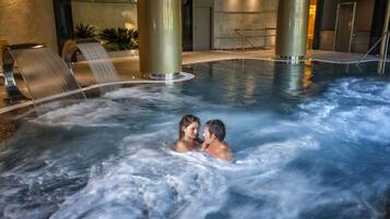 Una pareja en un jacuzzi con chorros de agua, rodeada de un moderno espacio interior con piscina que incluye columnas grandes y una cascada.