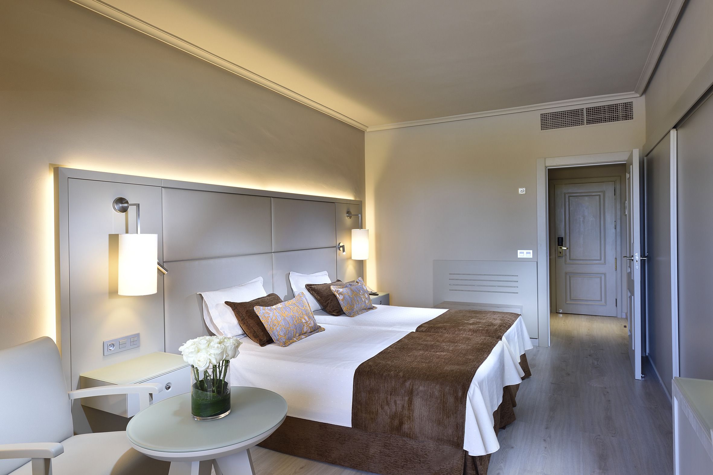 Una habitación de hotel con una cama grande, mesitas de noche, una pequeña mesa con un jarrón de flores y una puerta que da a otra habitación.