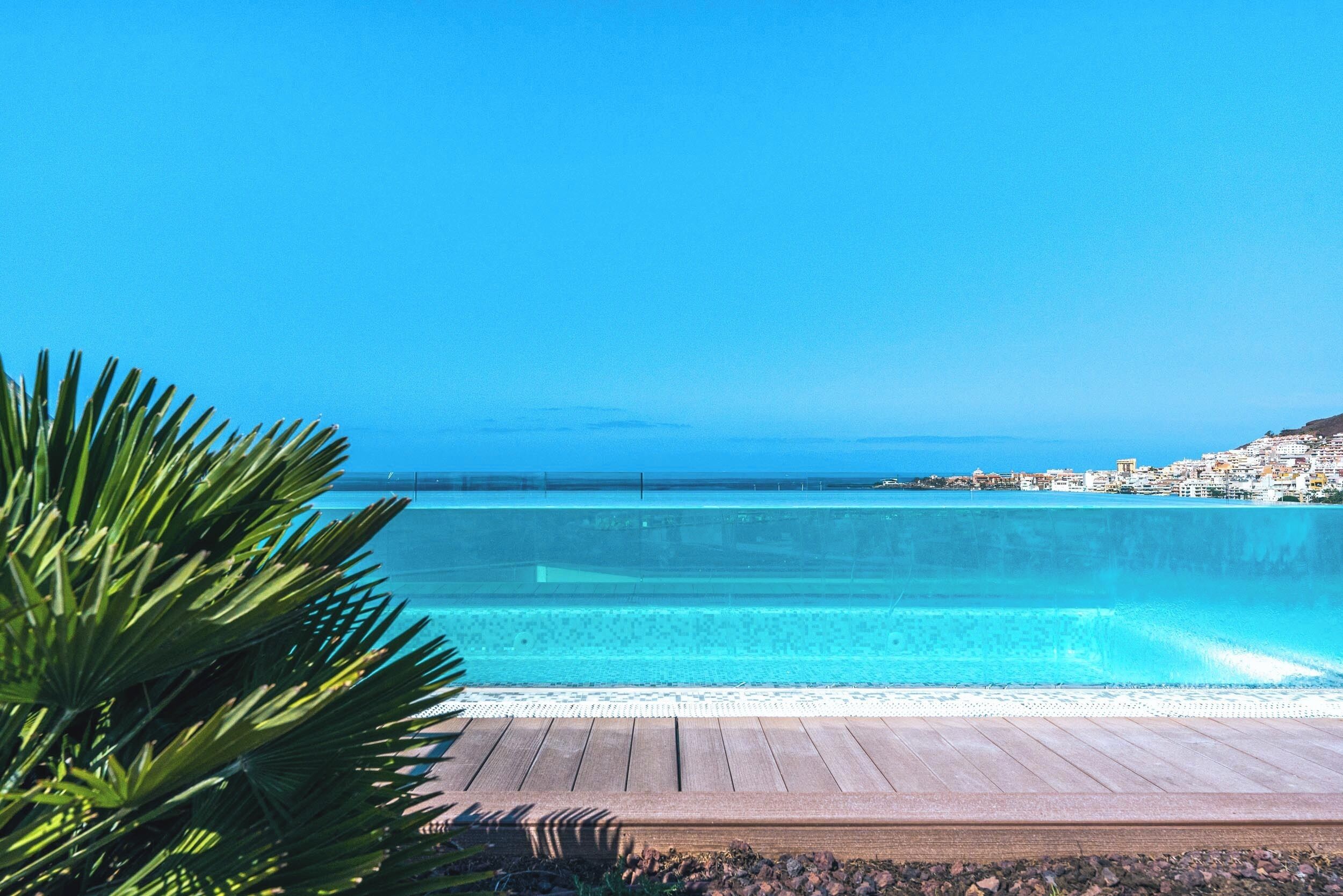 Cielo azul despejado, terraza de madera y piscina con vistas al océano y a un pueblo costero.