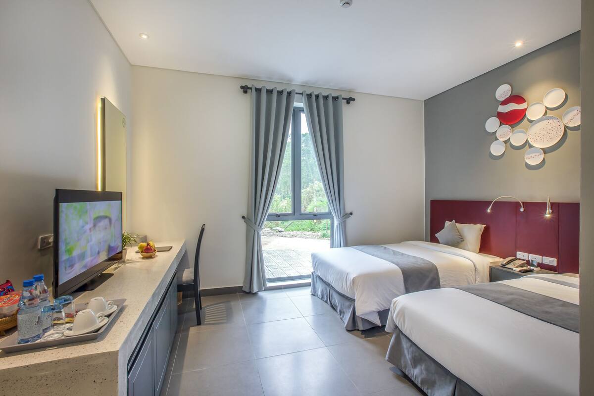 Habitación de hotel con dos camas, televisión, escritorio y un juego de botellas de agua, además de vistas al exterior.