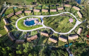 Vista aérea de un centro de bienestar con una piscina central, rodeada de cabañas con techo de paja y un sendero sinuoso.