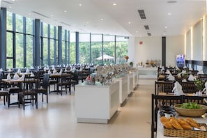 A spacious dining area with multiple tables and chairs, a buffet station, and large windows offering a view of greenery outside.