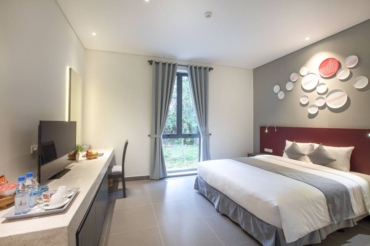 Habitación de hotel moderna con cama, televisor, escritorio con productos para el desayuno y un amplio ventanal con cortinas.