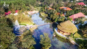 Vue aérienne d’un complexe hôtelier axé sur le bien-être, avec un étang central, un bâtiment circulaire surmonté d’un toit de chaume et une végétation luxuriante tout autour.