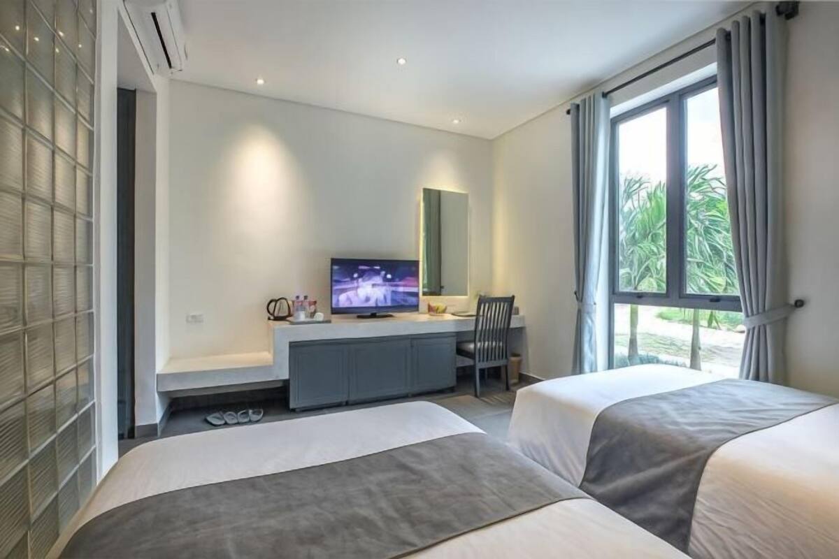 Habitación de hotel con dos camas, televisión, escritorio y ventana con vistas a la vegetación.
