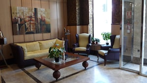 Un hall con un sofá, dos sillones, una mesa de centro con plantas, un cuadro en la pared y una puerta de cristal con letrero de salida.