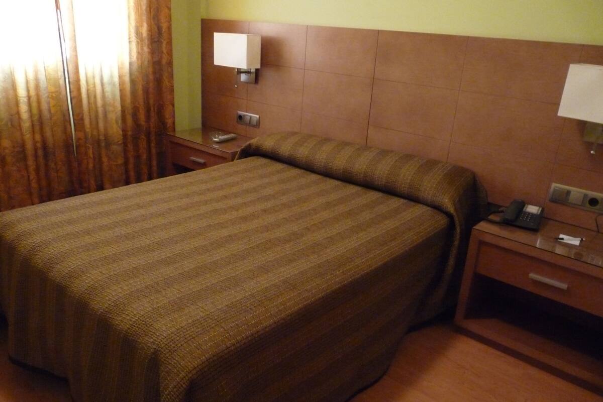 A hotel room with a large bed, a bedside table with a lamp, a phone, and a small drawer.