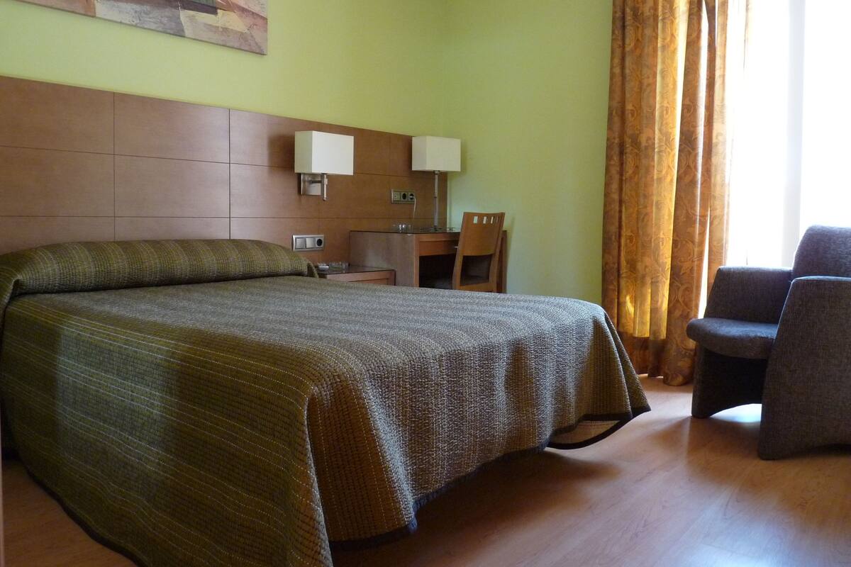 A hotel room with a large bed, a desk, a chair, and a window with curtains.