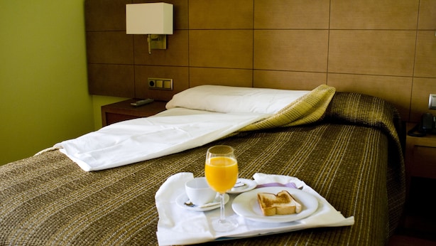 Una cama bien tendida con una bandeja de desayuno que incluye tostadas, un vaso de zumo de naranja y una taza.