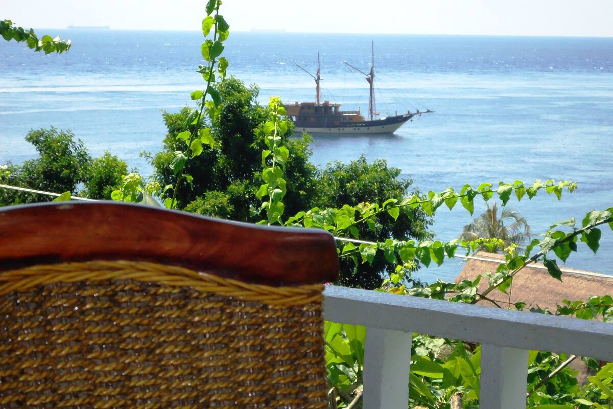 A view of a coastal area with a boat on the water, greenery, and a wicker chair.