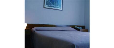 Una cama bien hecha con cabecera, una mesita de noche con lámpara, y un cuadro enmarcado en la pared.
