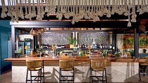 Un área de bar con tres taburetes de mimbre, una barra de madera y varias botellas y plantas.
