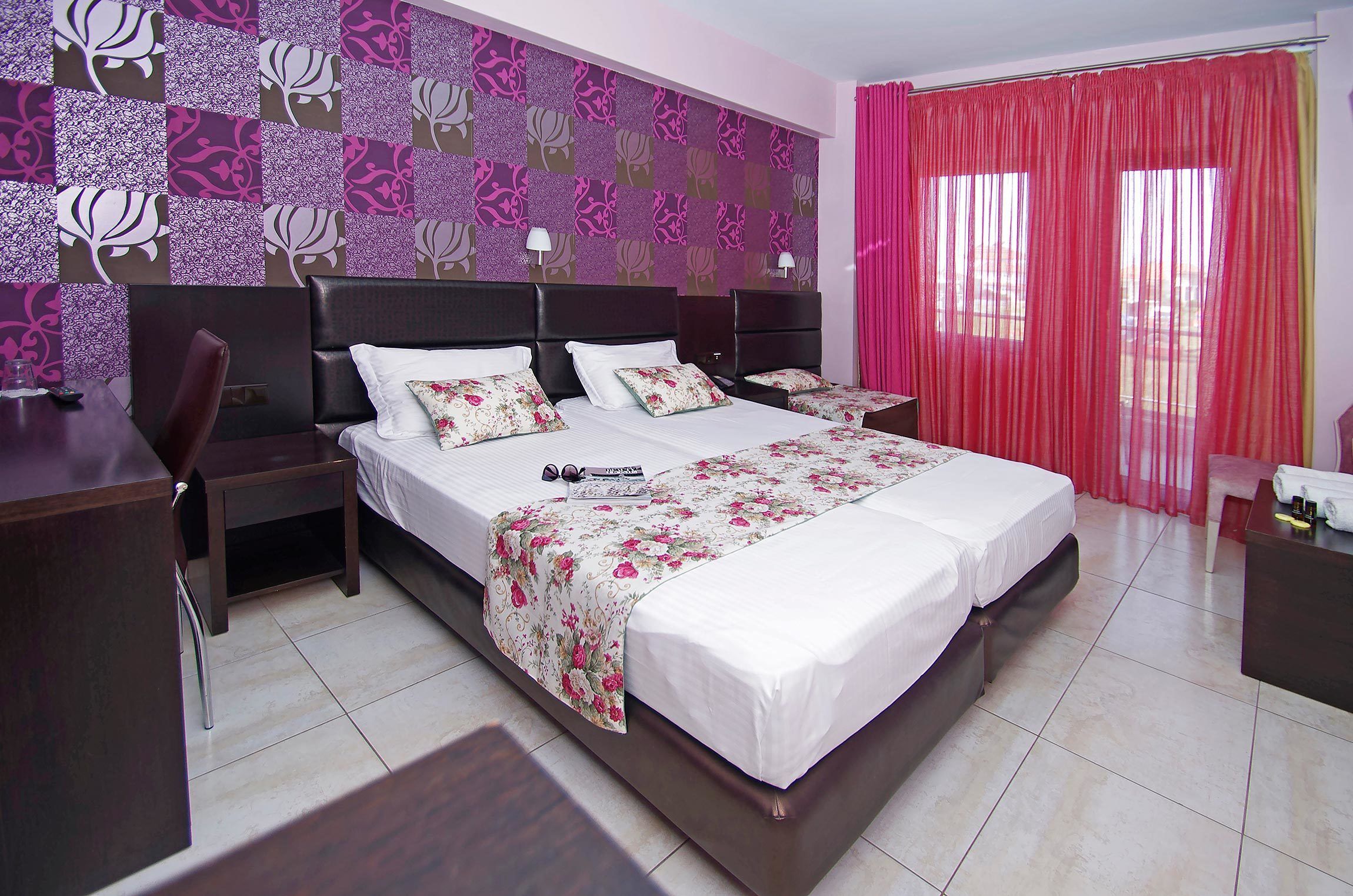 Una cama doble con una colcha estampada, dos mesitas de noche con lámparas, un ventanal con cortinas rojas y una pared con diseño.