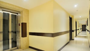 A hotel corridor with a glass elevator, a beige wall with a black stripe, and a tiled floor. - Hotel Aura, IGI Airport (New Delhi)