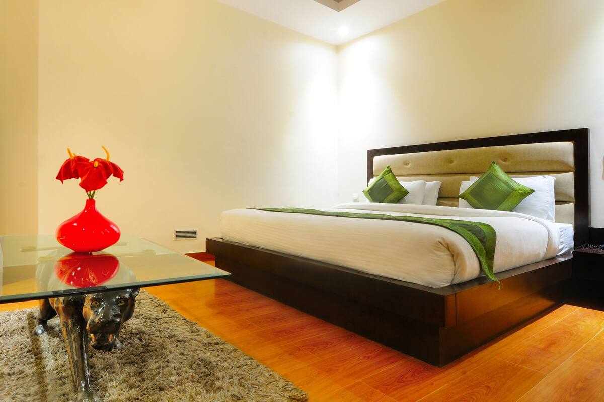 A modern bedroom with a glass coffee table, a red vase with flowers, a bed with green pillows, and a wooden floor.
