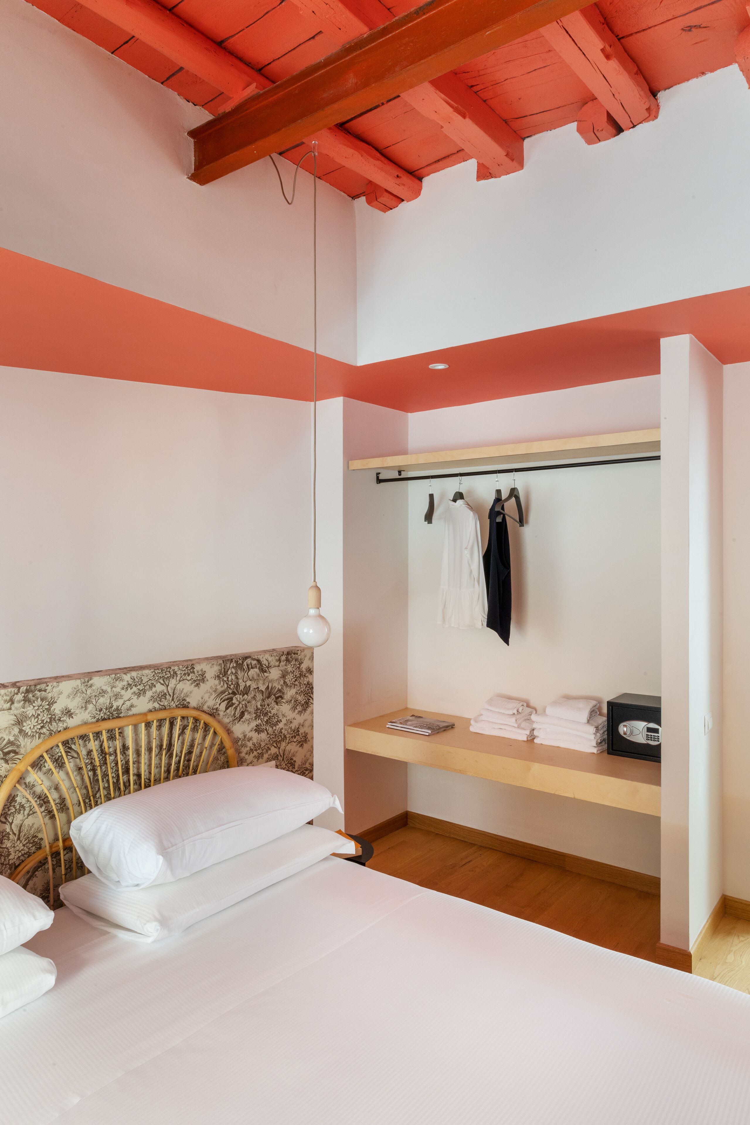Een slaapkamer met een bed, een houten hoofdbord, een hanglamp, een plank met opgevouwen handdoeken, en een kapstok met kleding.