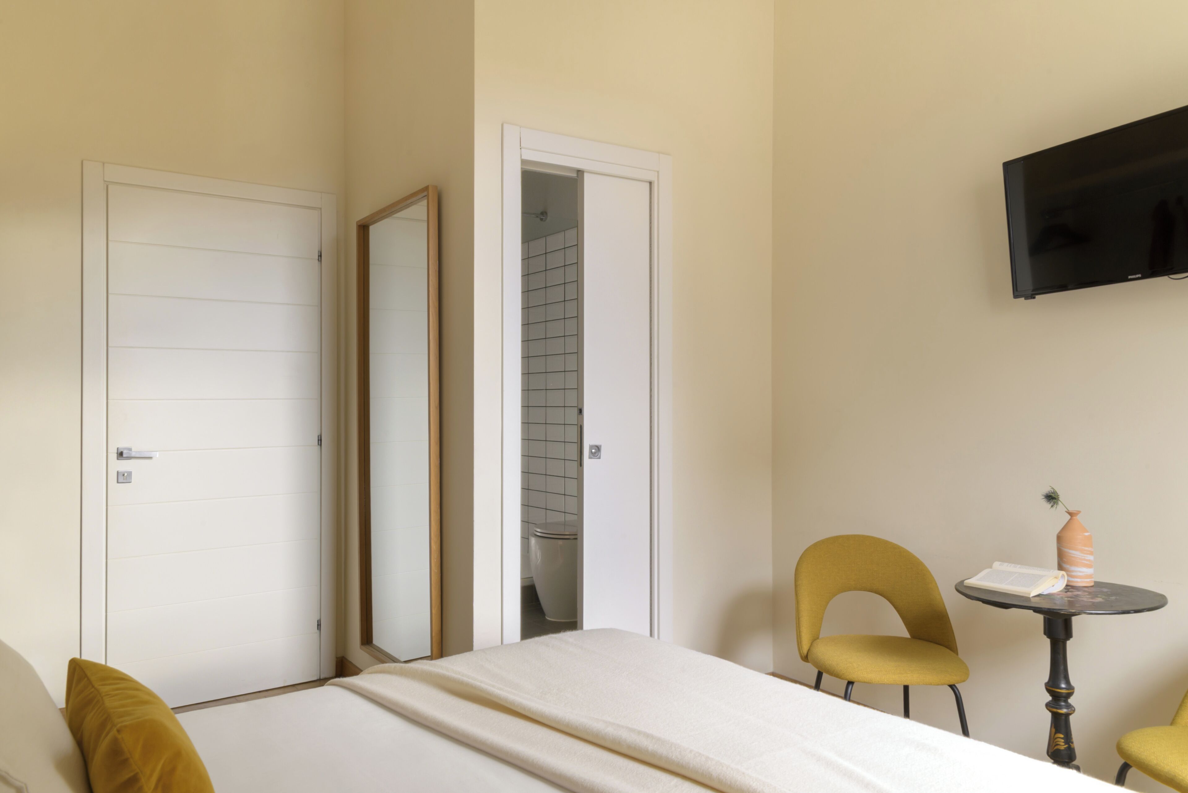 A hotel room with a bed, a chair, a small table with a book and a vase, a TV mounted on the wall, and a door leading to a bathroom.