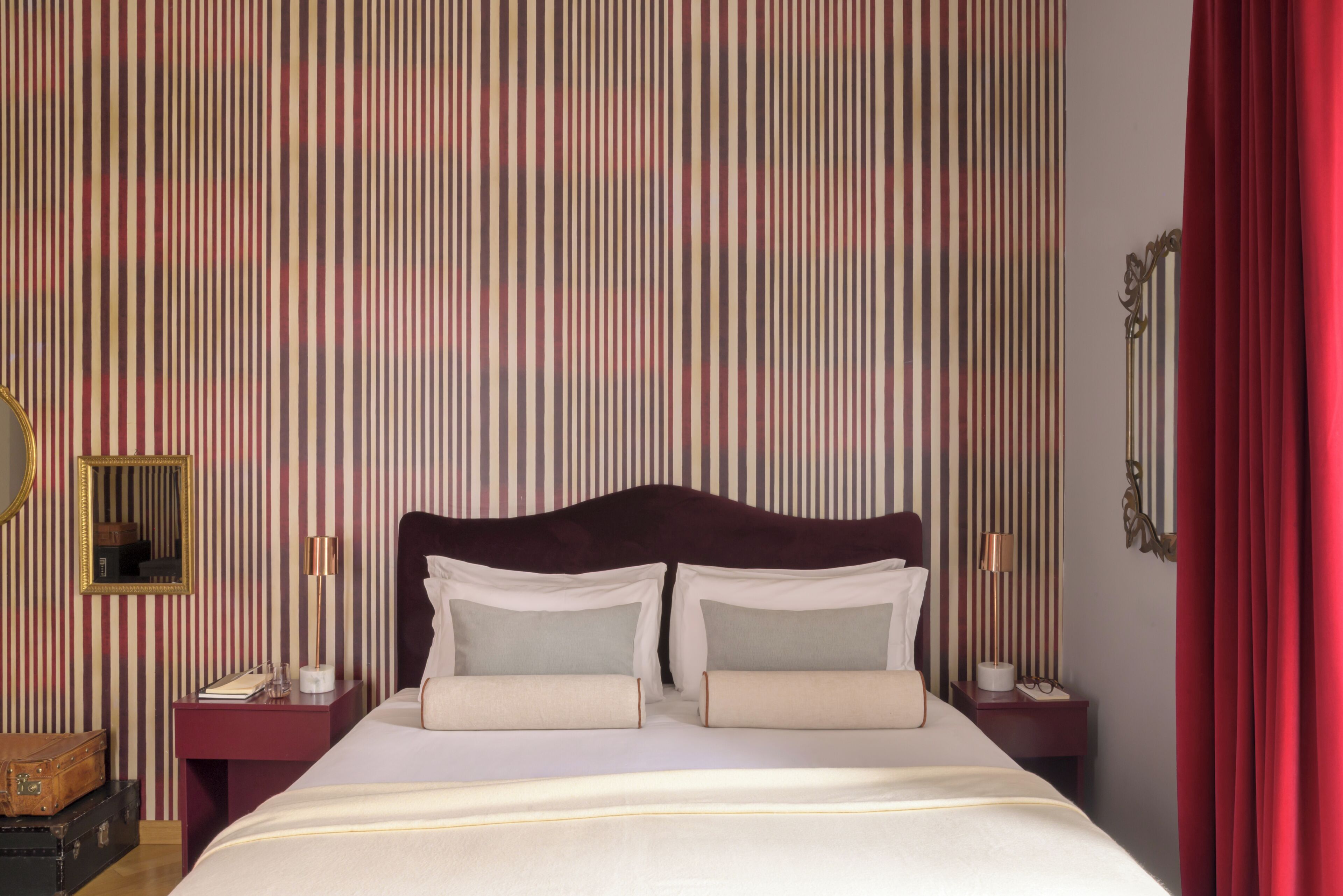 Habitación de hotel con papel tapiz a rayas, una cama grande con ropa blanca, dos mesitas de noche con lámparas y una cortina roja.