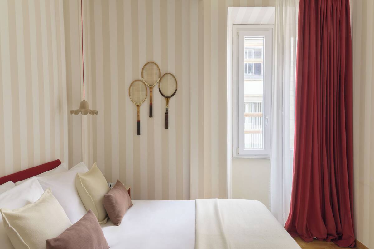 Ein Hotelzimmer mit gestreifter Tapete, einem Bett mit Kissen, einem Fenster mit Vorhängen und drei Tennisracket an der Wand.