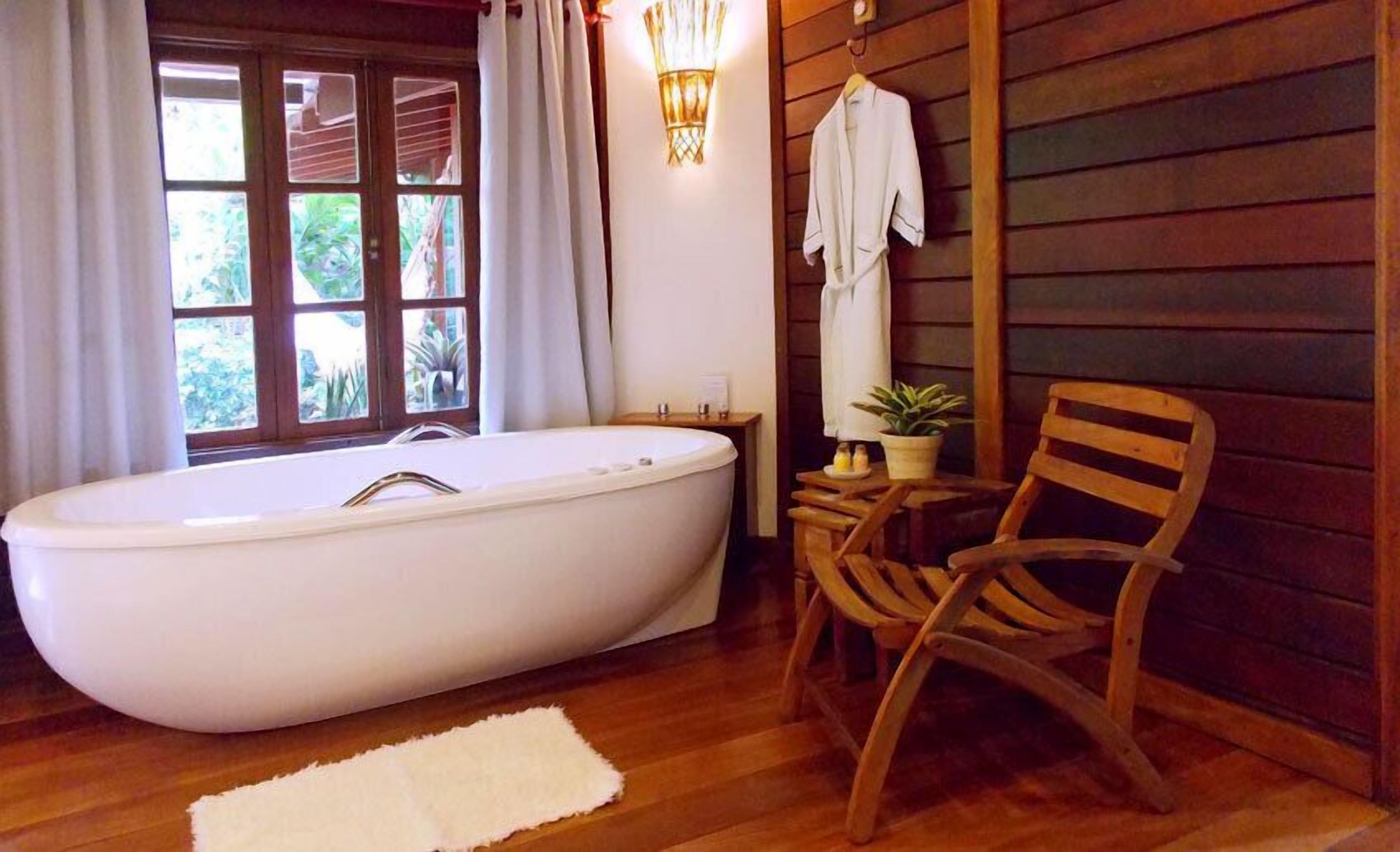 A bathroom with a freestanding bathtub, wooden flooring, a wooden chair, a small table with a plant, and a window with curtains.