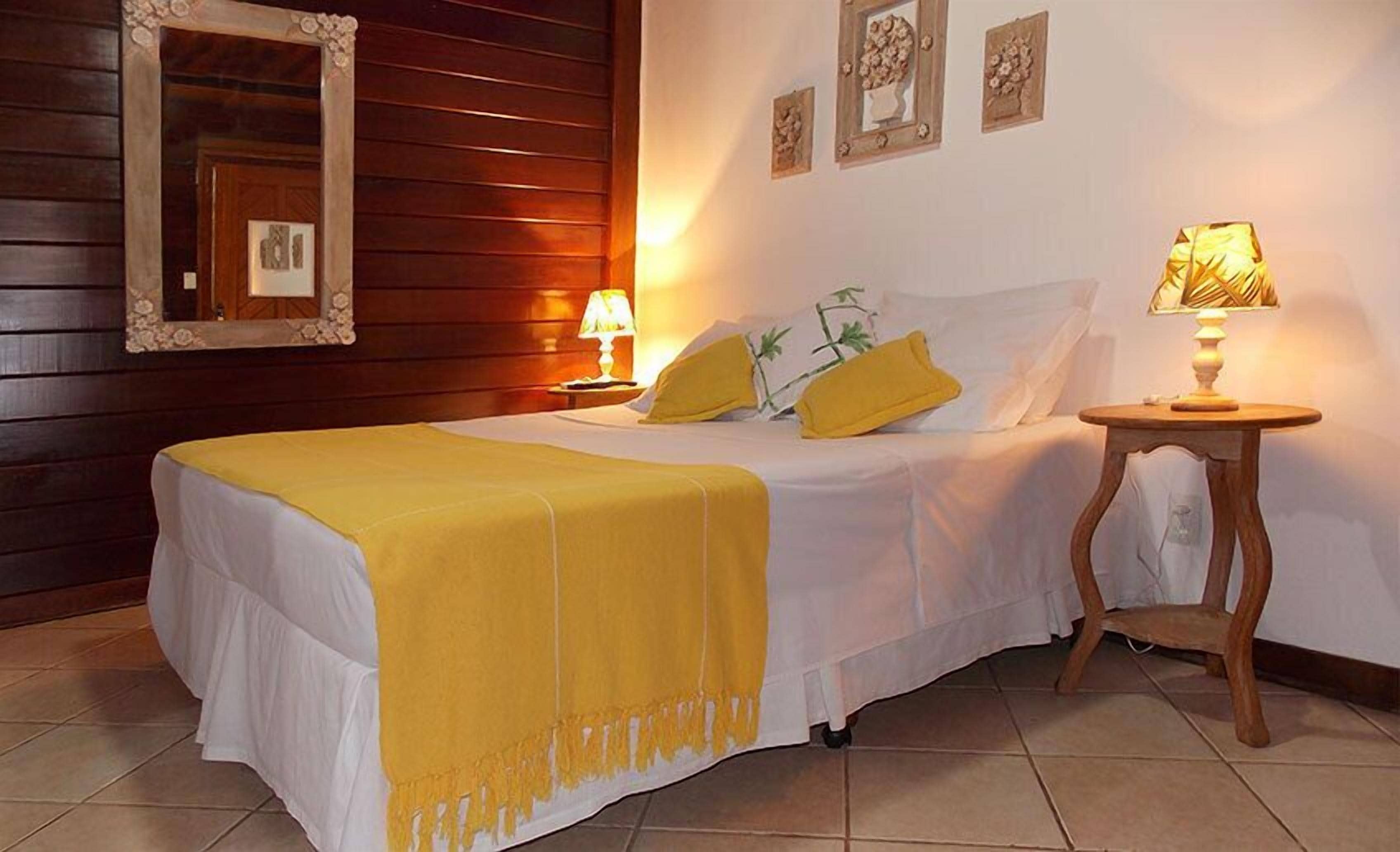 A bed with a yellow blanket, two pillows, a bedside table with a lamp, and a mirror on the wall.