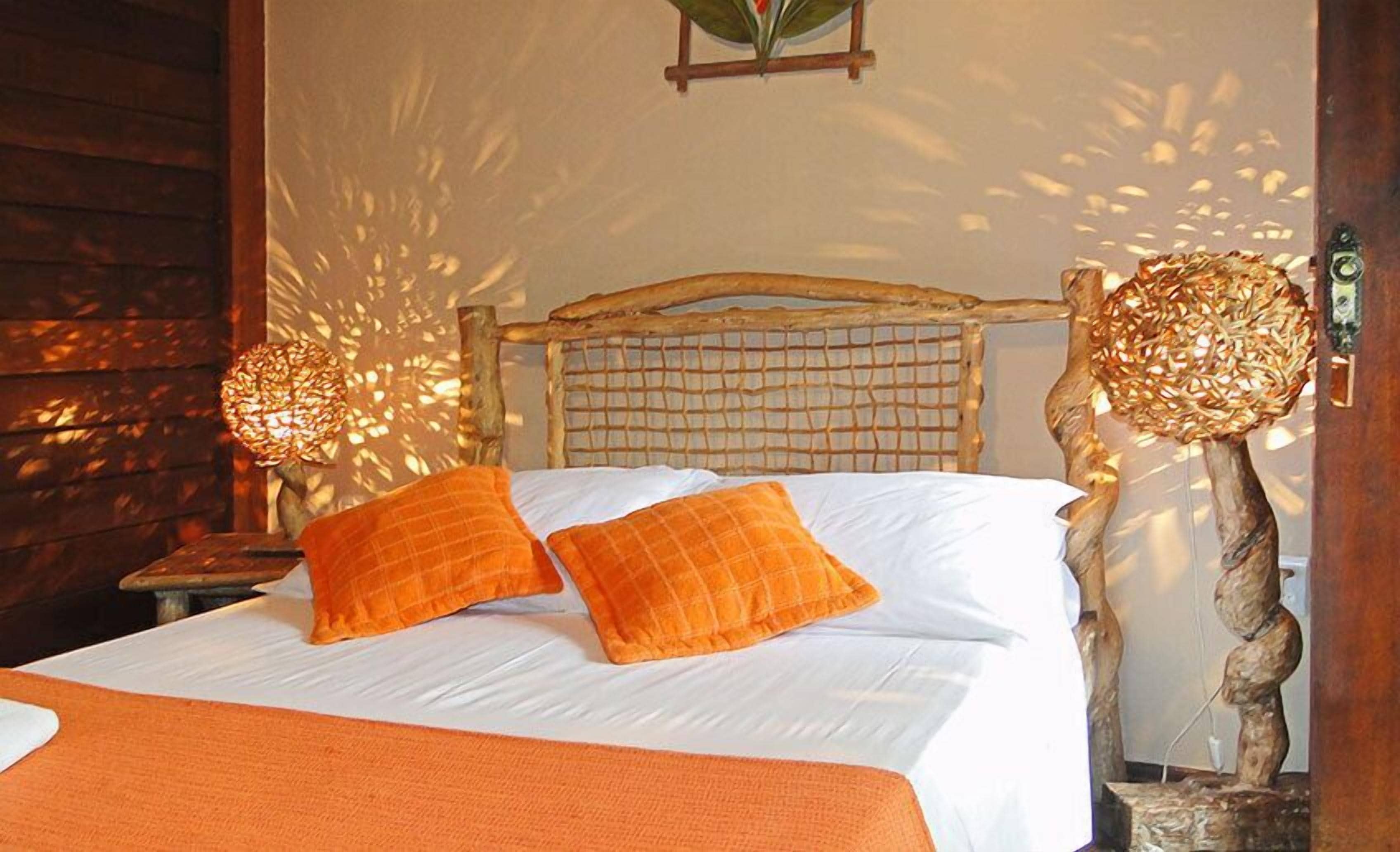 A bed with white linens and orange pillows, a wooden headboard, and two wall-mounted lamps with woven shades.