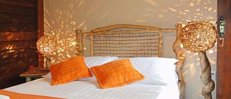 A bed with white linens and orange pillows, a wooden headboard, and two wall-mounted lamps with woven shades.
