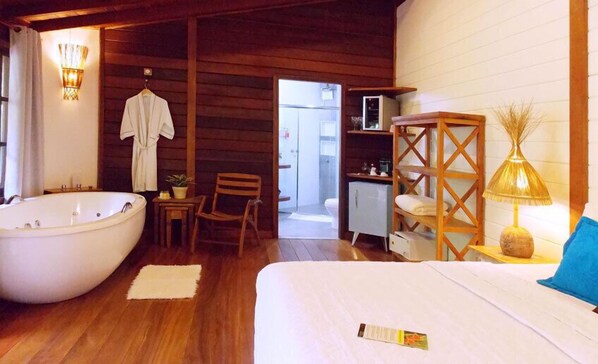A wooden cabin-style room with a large bathtub, a bed, a chair, a small table, a lamp, and a shelf with towels. - Pousada Riacho dos Cambucás (Angra dos Reis)