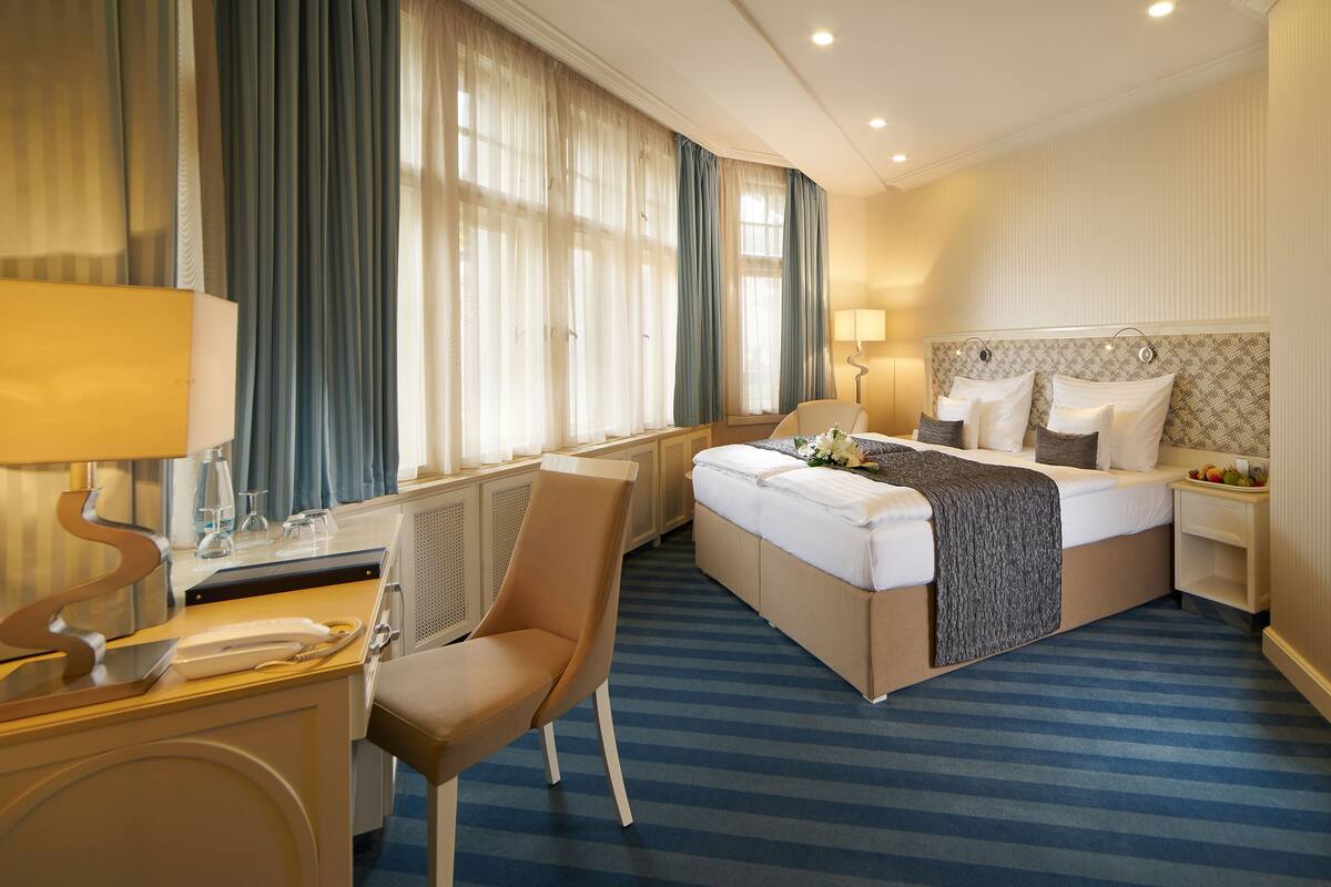 Ein Hotelzimmer mit Bett, Schreibtisch, Stuhl, Lampe und einem fenster mit Vorhängen.