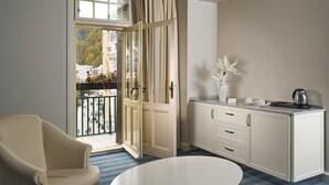 Een kamer met een groot raam, een stoel, een eettafel en een keukenhoek met kastjes en een waterkoker.