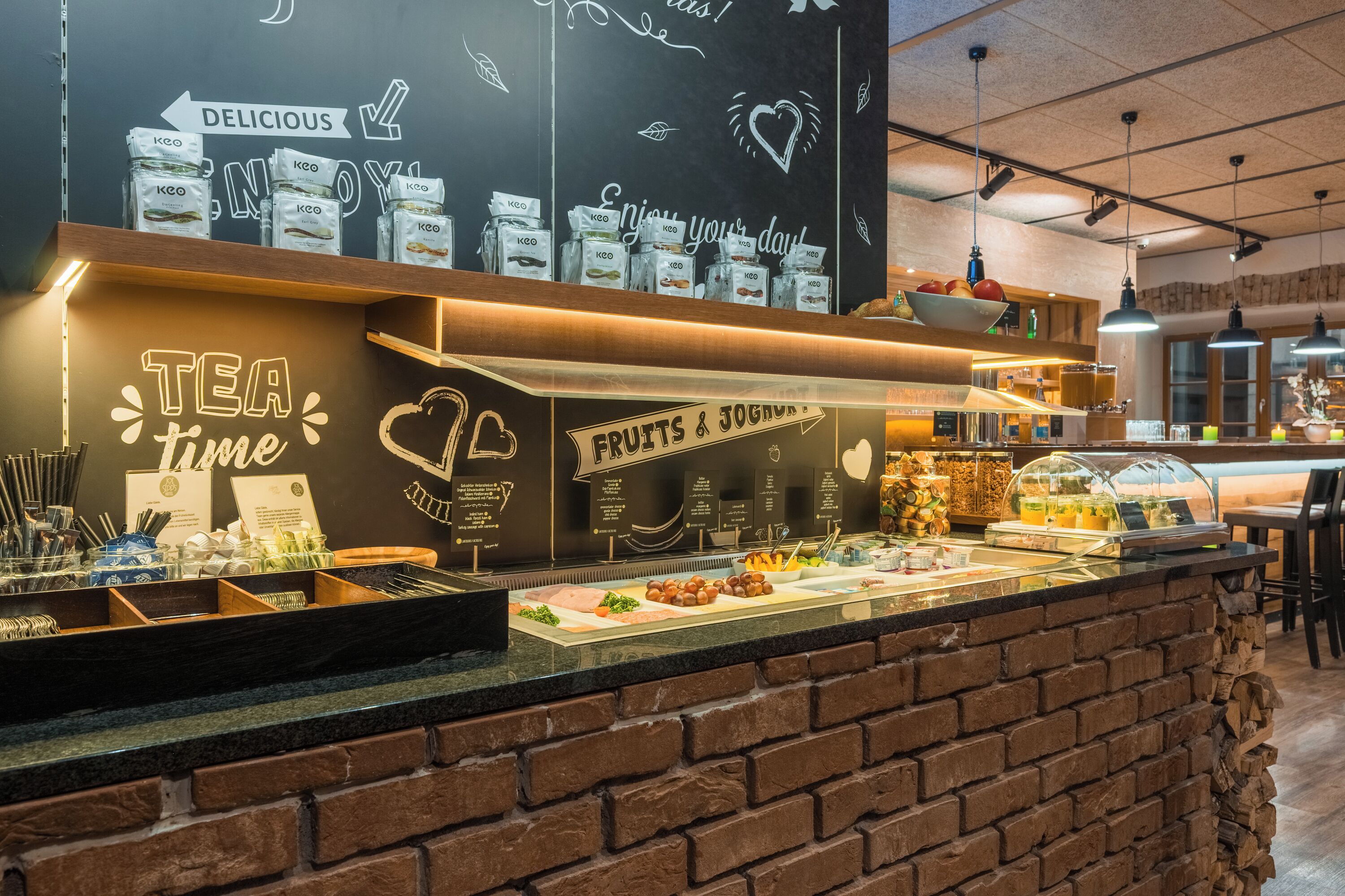A coffee shop counter with a chalkboard menu, tea and fruit display, and a brick wall design.