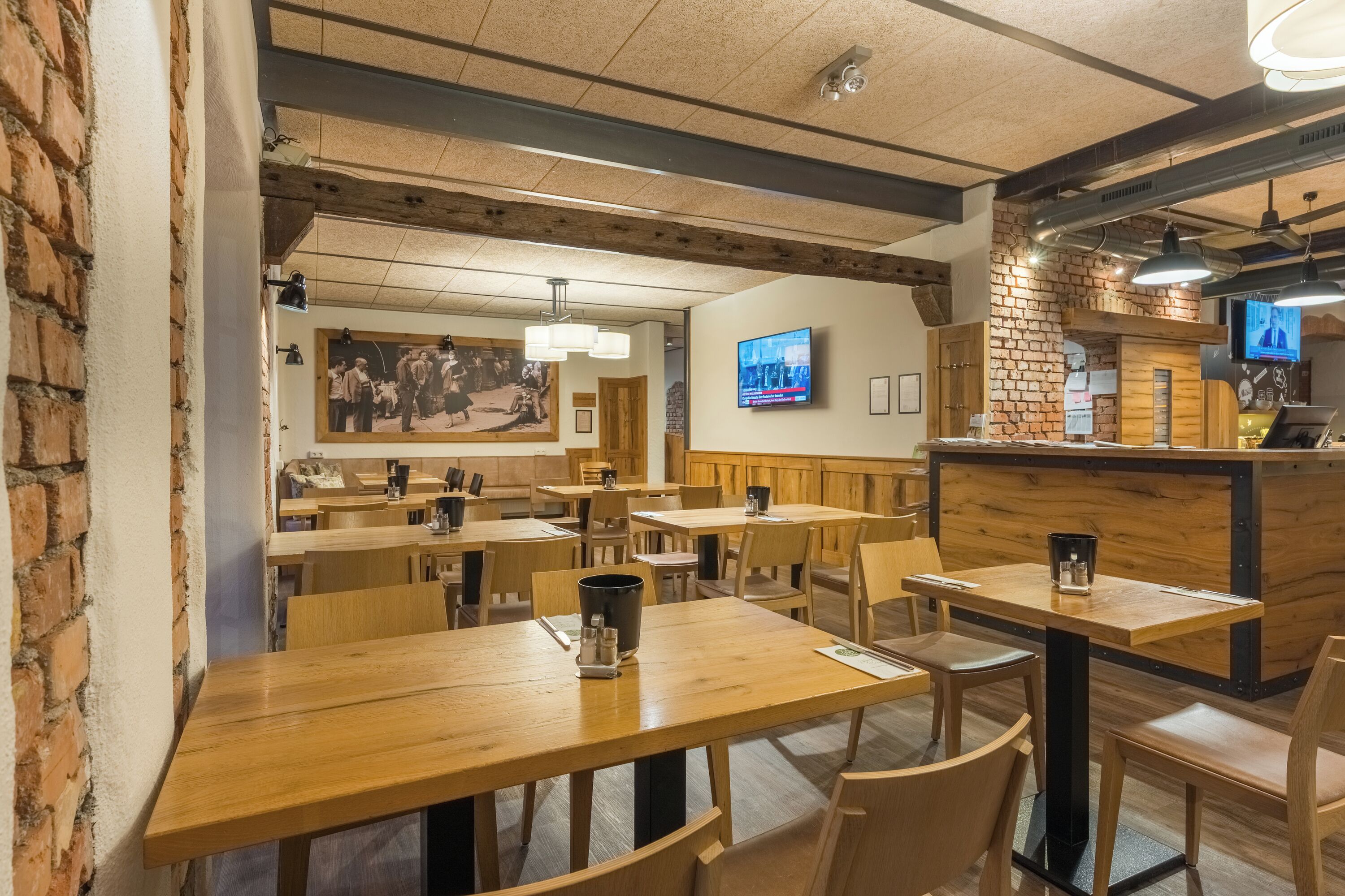 A restaurant interior with wooden tables and chairs, a brick wall, a television mounted on the wall, and a ceiling with exposed beams.