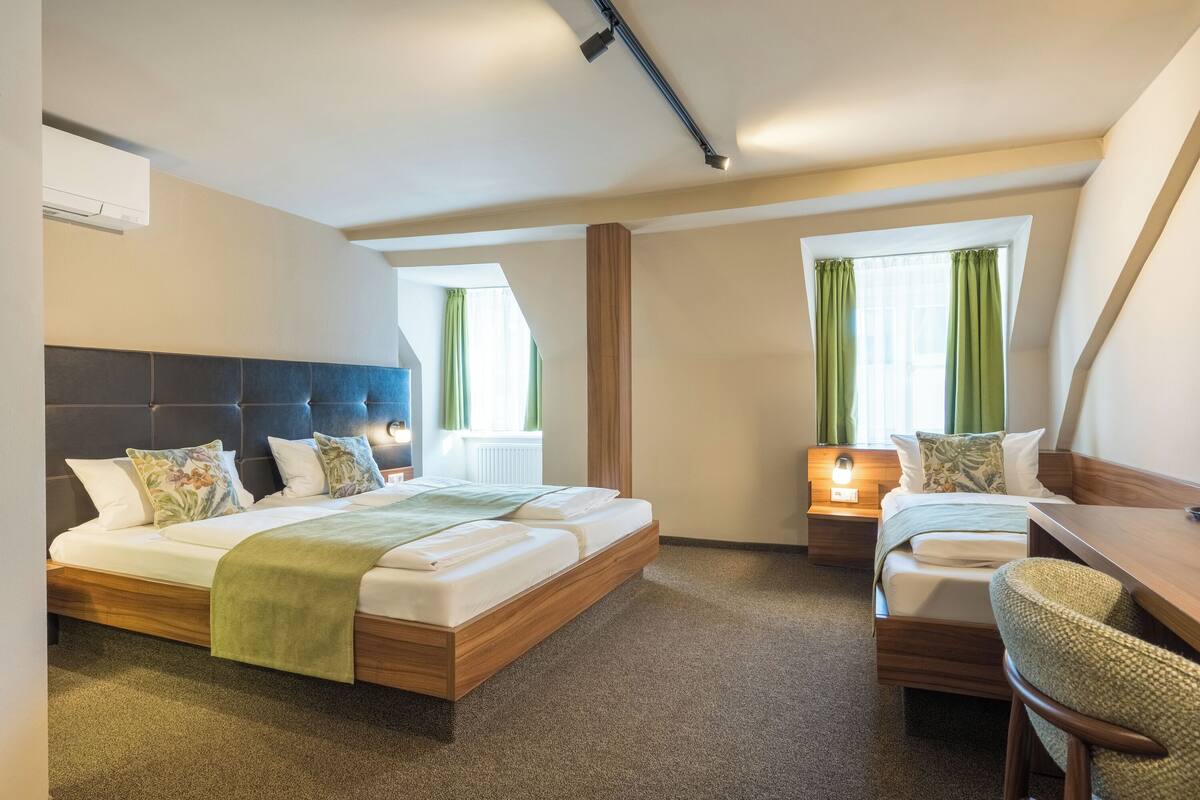 Een hotelkamer met een groot bed, een bureau en een stoel. Twee ramen met gordijnen, een nachtkastje en een wandairconditioner.