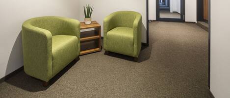 A hallway with two green armchairs, a small wooden table with a plant, and a door with an emergency exit sign.