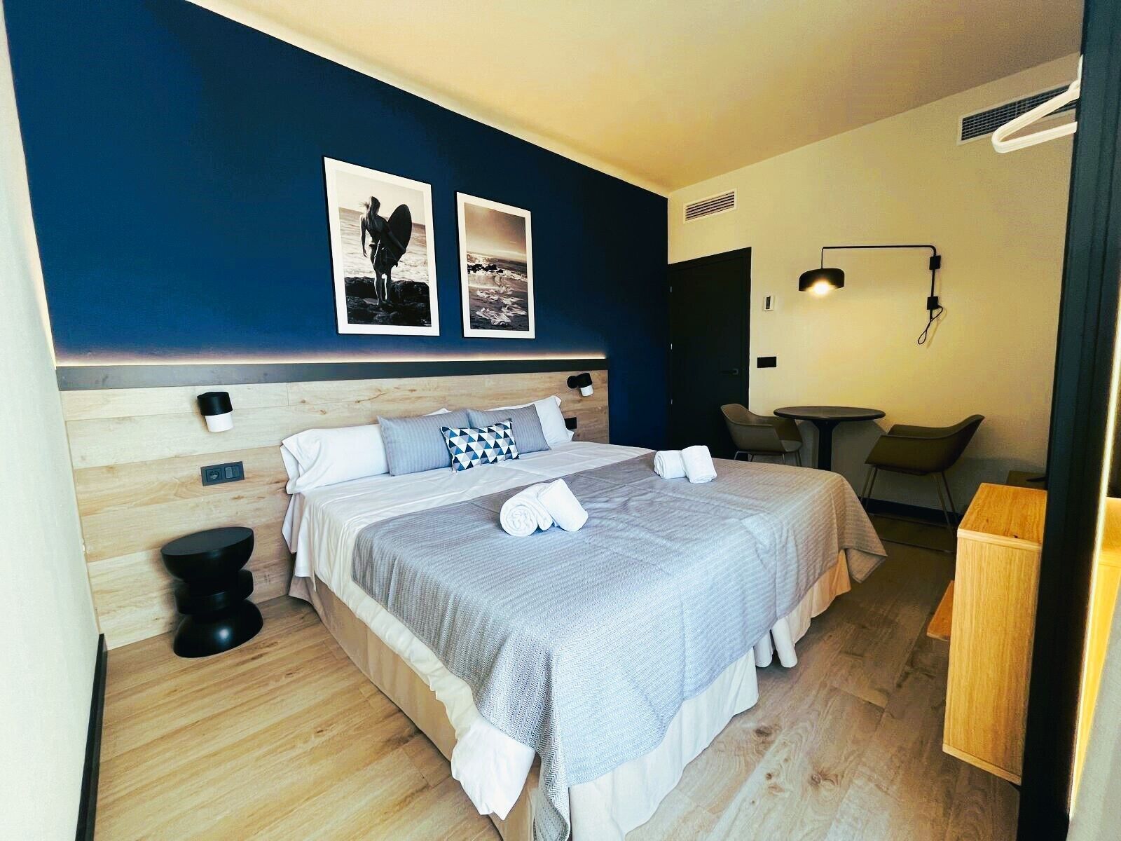 Una habitación de hotel moderna con una cama grande, una mesita de noche, una silla y un pequeño escritorio. Hay dos cuadros enmarcados en la pared sobre la cama.