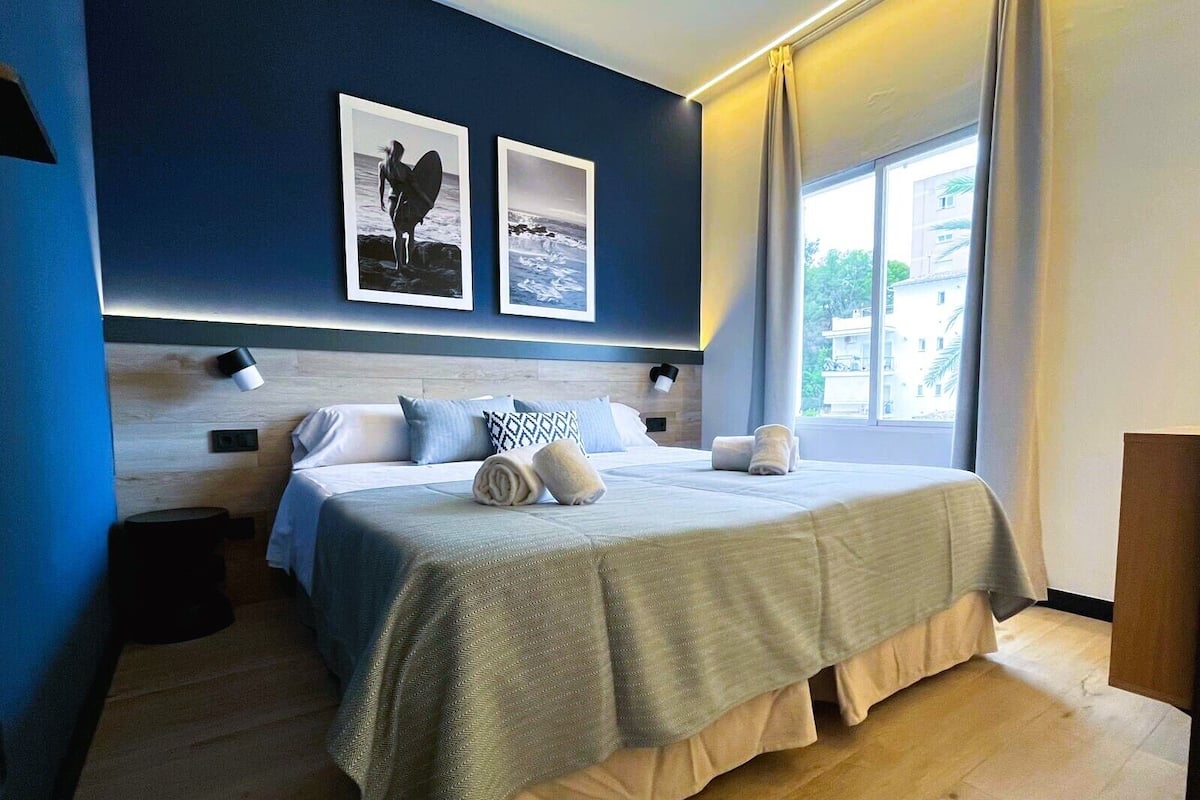 Une chambre d’hôtel moderne avec un grand lit, une tête de lit en bois, deux tableaux encadrés au mur, une lampe de chevet et une vue sur l’extérieur par une fenêtre.