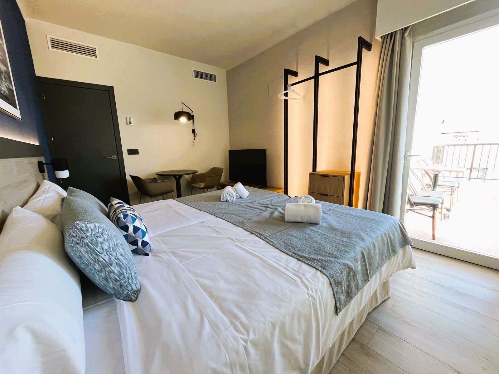 A hotel room with a large bed, two bedside tables, a desk, a chair, a TV, and a balcony with outdoor seating.