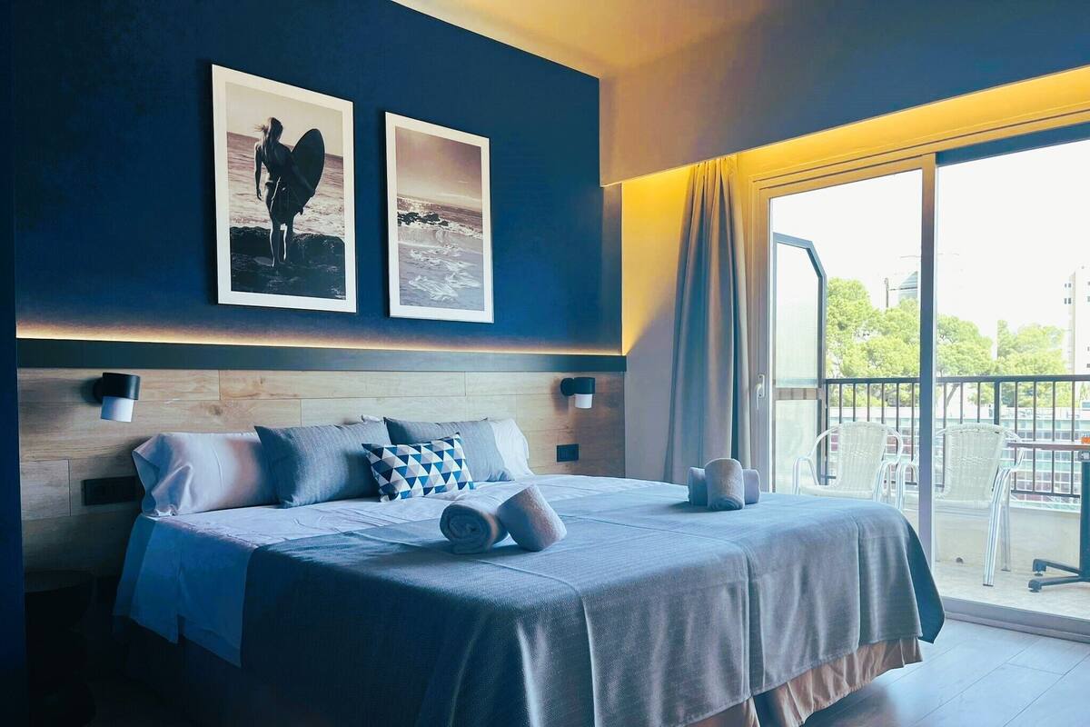 Une chambre moderne avec un grand lit, deux cadres photos accrochés au mur, un balcon avec des chaises et une vue sur la ville.