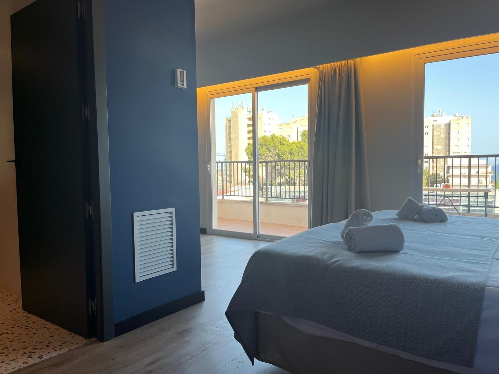 Una camera d'hotel con un grande letto, una porta scorrevole in vetro che dà su un balcone e vista su edifici e alberi.