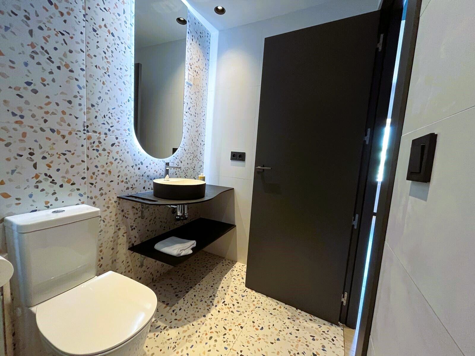 A modern bathroom with a round sink, a toilet, and a dark door.
