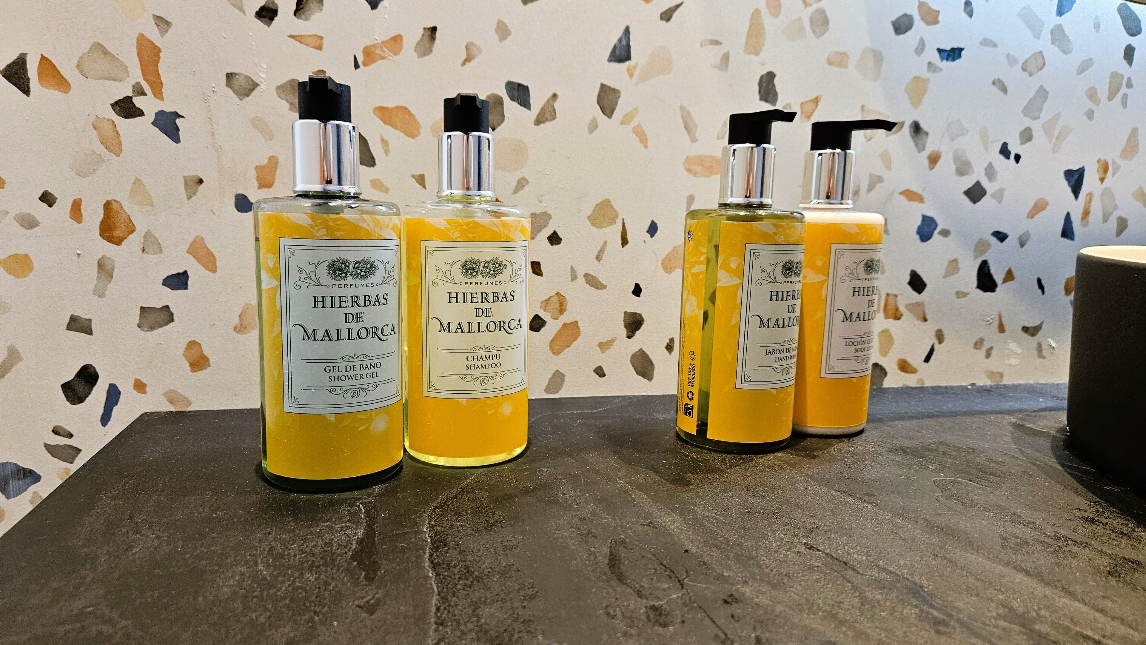 Three bottles of gel de baño on a wooden surface against a terrazzo wall.